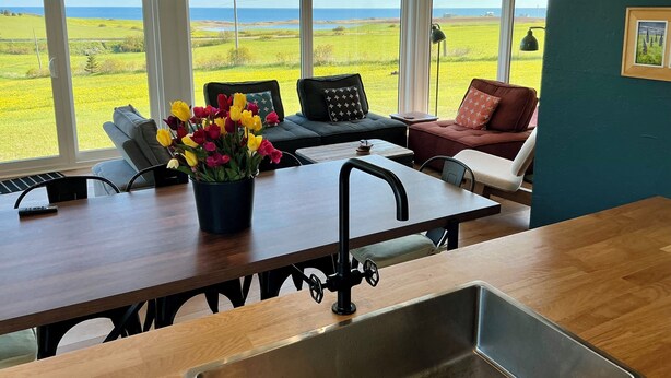 A kitchen with a view of the ocean, a dining table, and a sofa with a tulip vase. - Country cottage, exceptional view, lake, dunes and sea. 5 minutes from the beach (Kensington)