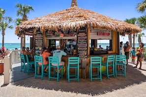 A beachside bar with a thatched roof, outdoor seating, and a menu board.