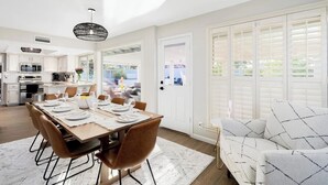 A modern kitchen with white cabinetry, a dining area with a wooden table set for four, and a large window with blinds.