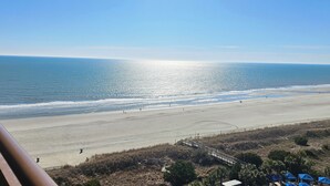 Een strand met helder uitzicht op de oceaan, een zandstrand en enkele mensen die er wandelen. Er is een houten looppad naar het strand en enkele gebouwen met blauwe daken op de voorgrond.