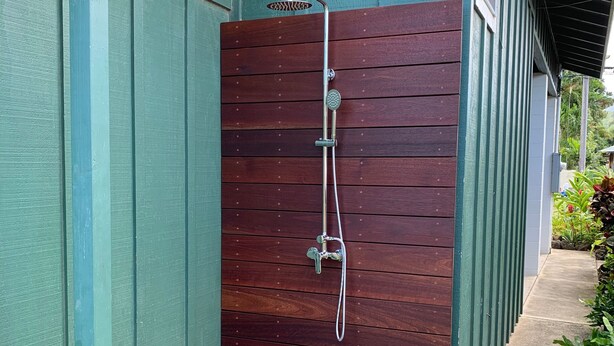 Un cerramiento de ducha de madera con cabezal y grifo de metal, junto a un edificio verde.