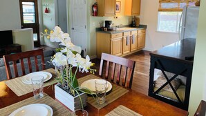 Un comedor con una mesa de madera dispuesta para cuatro, una cocina con armarios de madera y un refrigerador, y un ventilador de techo.