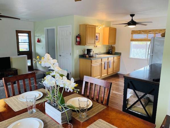 A dining area with a wooden table set for four, a kitchen with wooden cabinets and a refrigerator, and a ceiling fan.