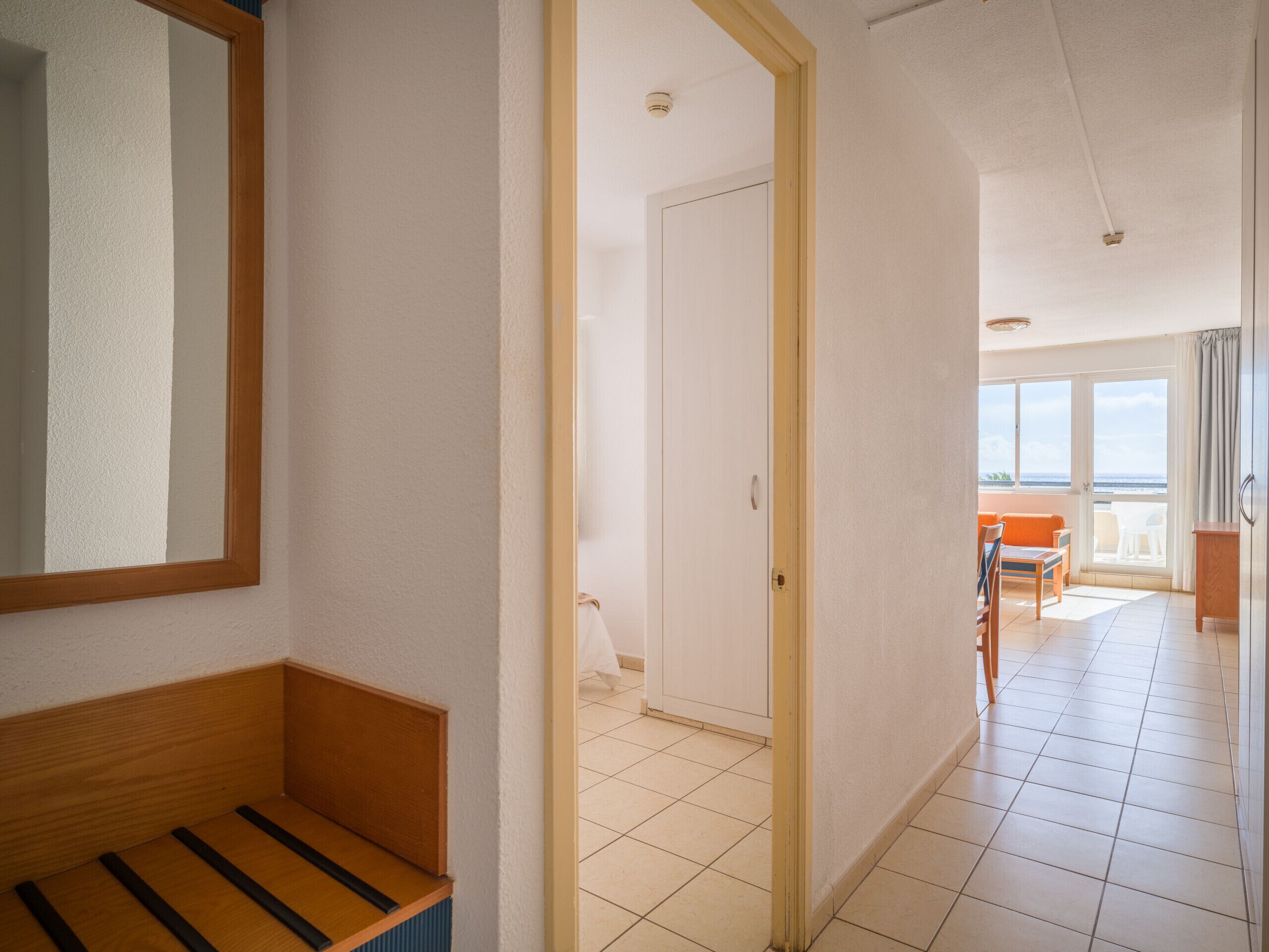 A hotel room with a wooden bench, a door leading to a balcony with a view of the sea, and a tiled floor.