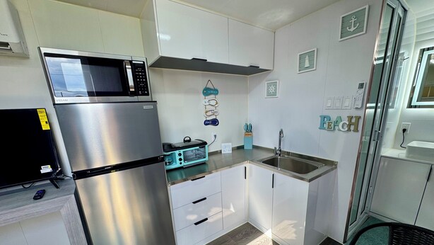 A compact kitchen with a stainless steel refrigerator, a microwave, and a toaster oven. There are white cabinets, a sink, and a small dining area with a glass table. - Rincon Sunset Studio (Rincon)