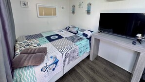 A compact room with a bed, a desk, a television, and a window with blinds. - Rincon Sunset Studio (Rincon)