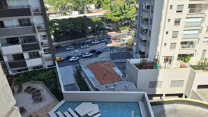 En pool på taget med fliseterrasse og liggestole, omgivet af høje bygninger og en parkeringsplads.