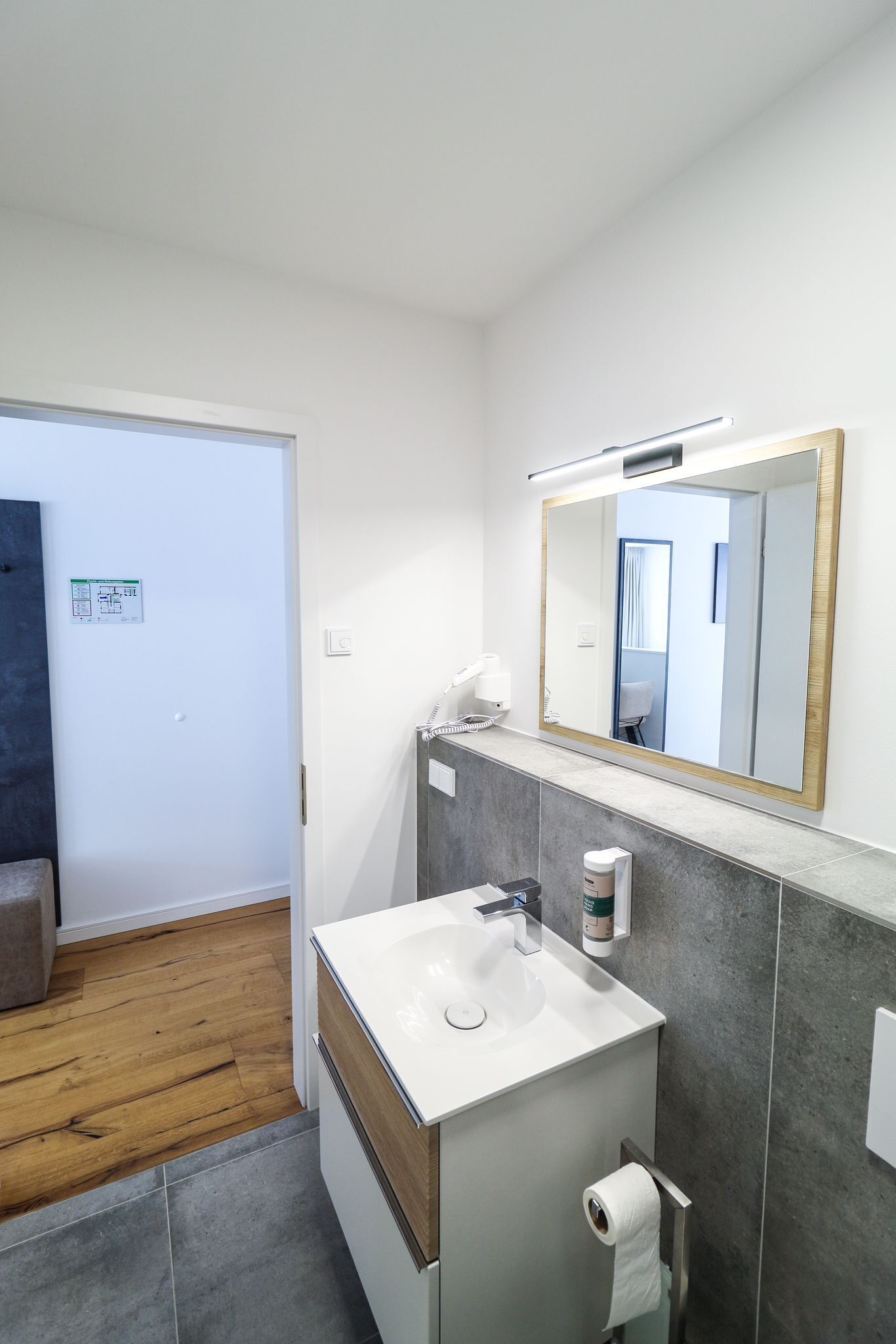 A modern bathroom with a white sink, a large mirror, and a towel dispenser.