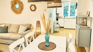 Un salon avec un canapé, une table à manger entourée de chaises, un luminaire suspendu et, au fond, une cuisine.