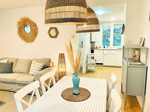 A living room with a sofa, a dining table with chairs, a hanging light fixture, and a kitchen in the background.
