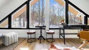 A modern loft bedroom with a sloped ceiling, large windows, a desk, two stools, and a cowhide rug.
