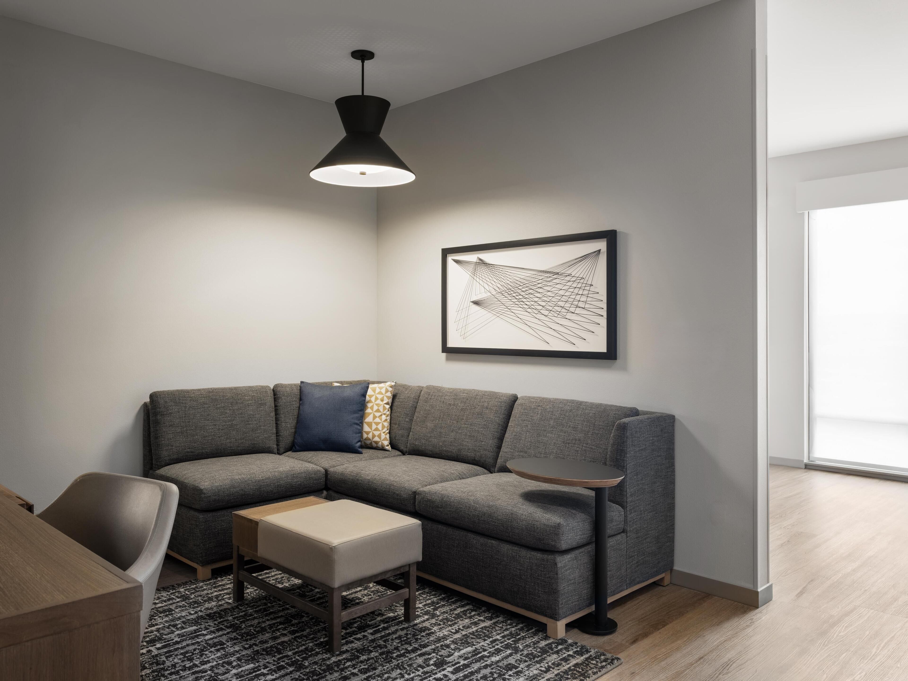 A modern living room with a gray sectional sofa, a small wooden coffee table, a black pendant light, and a framed abstract art piece on the wall.