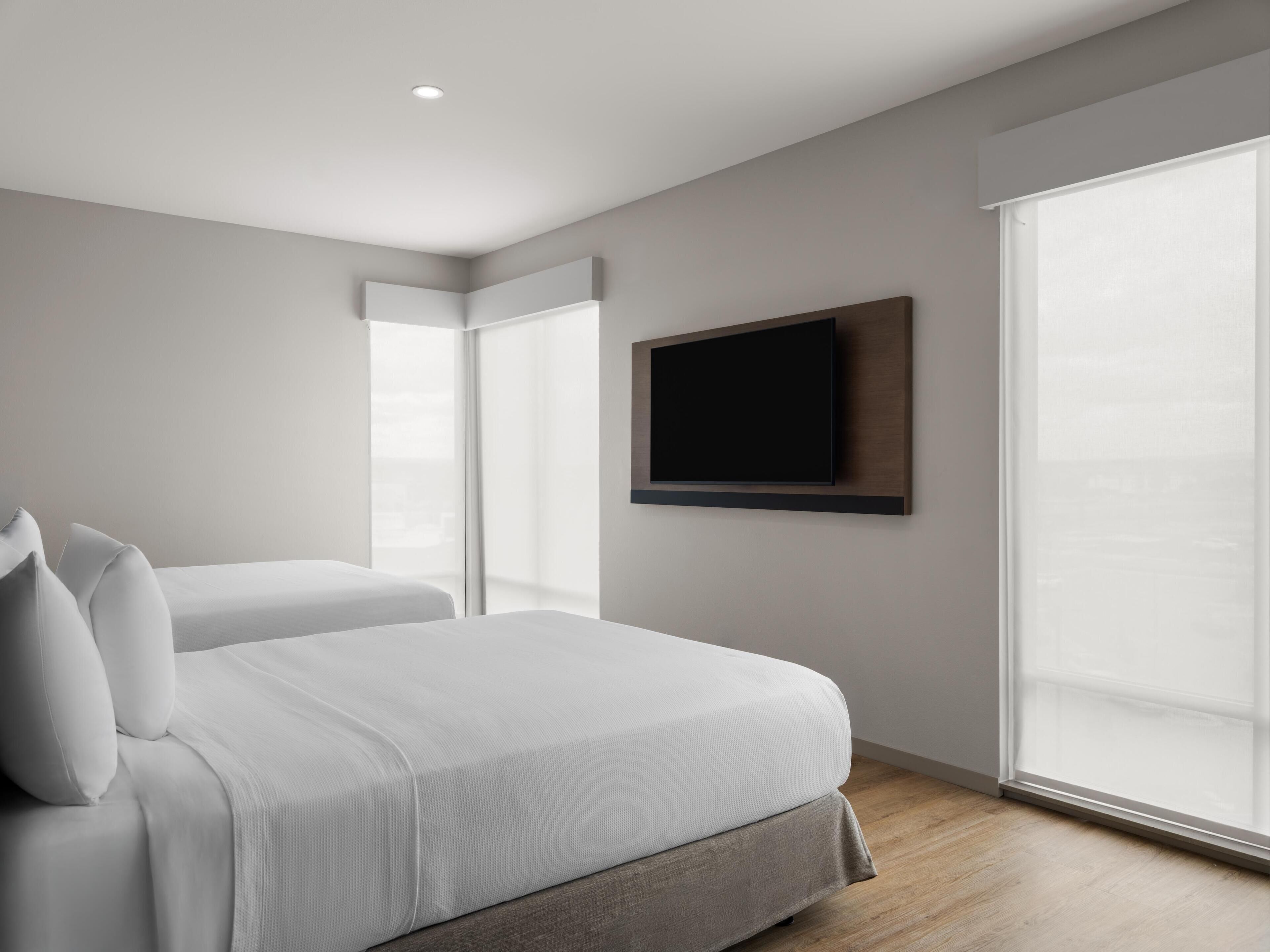 A modern hotel room with a large bed, a flat-screen TV mounted on the wall, and a large window providing natural light.