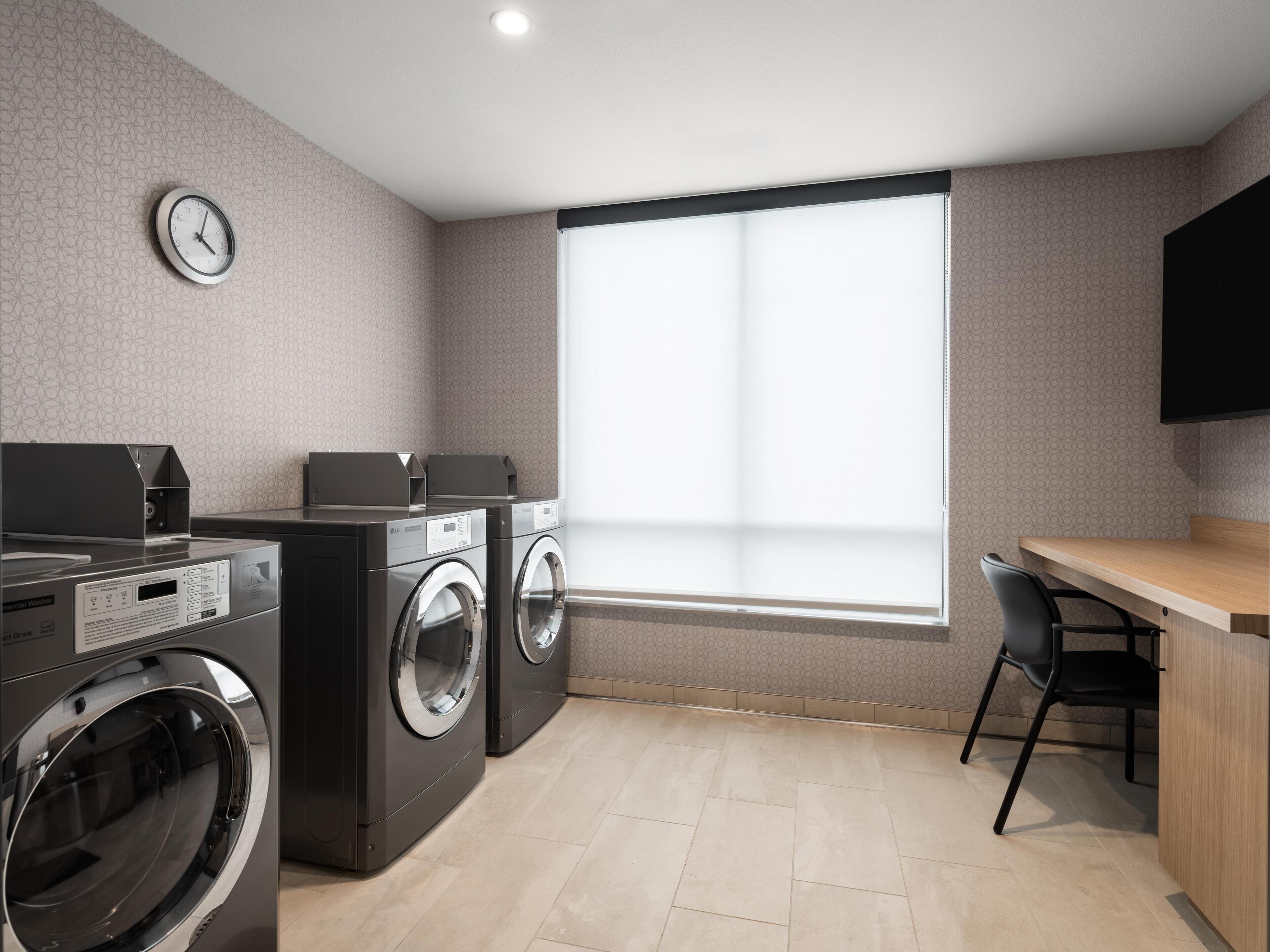 A laundry room with two front-loading washing machines, a wall-mounted clock, a large window, and a wooden desk with a chair.