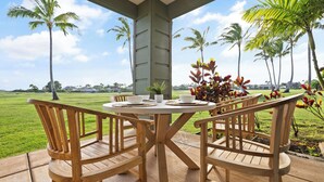 A wooden deck with a round table and chairs, surrounded by palm trees and a view of a green field.