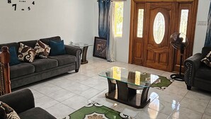 Ein geräumiges Wohnzimmer mit einer großen Couch, einem Glastisch und einem gemusterten Teppich. Eine Holztür und ein Fenster mit Vorhängen sind ebenfalls vorhanden.