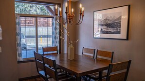 A dining area with a wooden table, six chairs, a chandelier, and a framed picture on the wall. - 2 Bedroom at Mystic Springs Chalets & Hot Pools (Canmore)