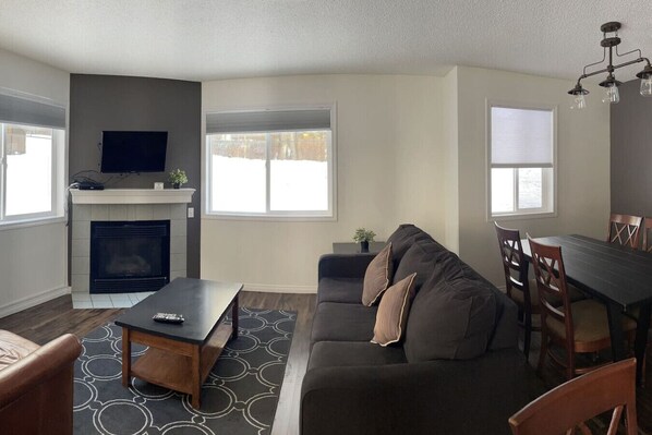 A living room with a fireplace, sofa, coffee table, TV, dining table, and chairs. - 2 Bedroom Condo at Canmore's Banff Boundary Lodge (Harvie Heights)