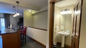A hotel room with a kitchen area, a dining table, and a bathroom with a sink and mirror.
