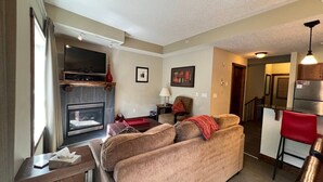 Ein Wohnzimmer mit einer Couch, einem Kamin, einem Fernseher und einer Küchenzeile im Hintergrund.