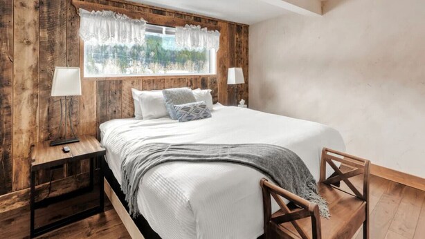Een slaapkamer met een houten hoofdbord, een groot bed, twee bedlampen, een raam met kantgordijnen en een houten stoel.