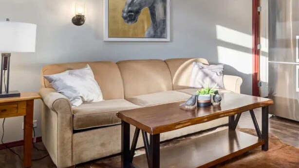 Ein Wohnzimmer mit einem beigefarbenen Sofa, einem Holztisch, einer Lampe und einem gerahmten Gemälde von Pferden an der Wand.