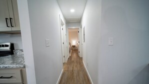 A narrow hallway with white walls and wooden flooring, leading to a room with a bed and a nightstand. - Charming 2-bedroom farmhouse in nice Wendell with AC, WiFi (Wendell)