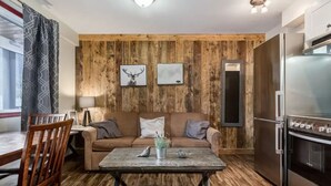 A cozy living room with a wooden wall, a sofa, a dining table, and a modern kitchen with a stainless steel refrigerator. - Yam 27/28 - Upper 2 Bedroom K/Q (Dead Man's Flats)