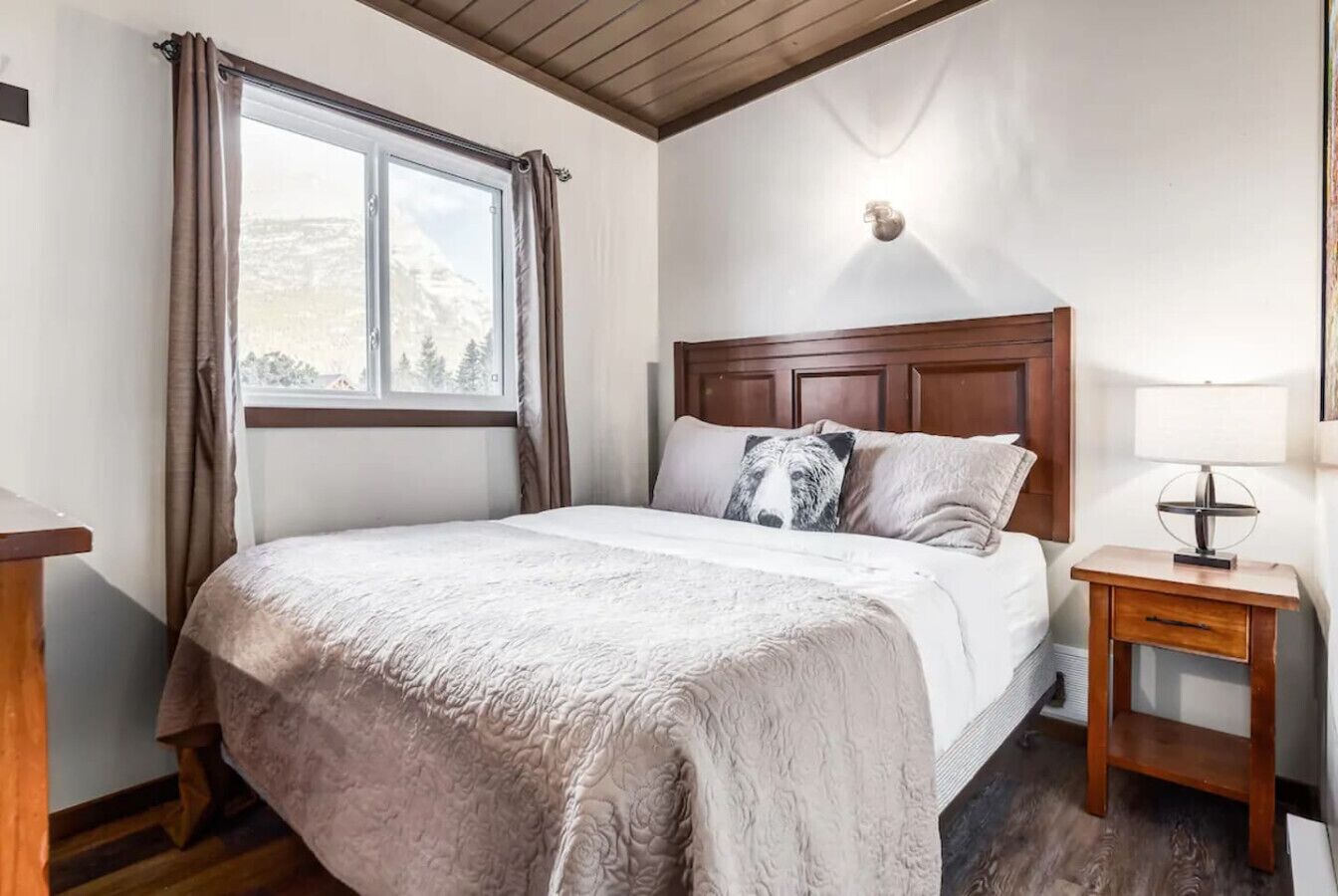 Un dormitorio con cama, ventana con cortinas, mesita de noche con lámpara, y una lámpara de pared.