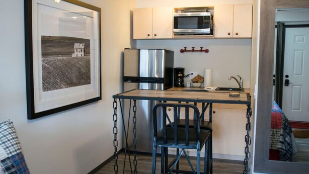 A compact kitchen with a microwave, refrigerator, and coffee maker. There is a framed picture on the wall and a small table with chairs. - Yam 12 - Studio with King (Dead Man's Flats)