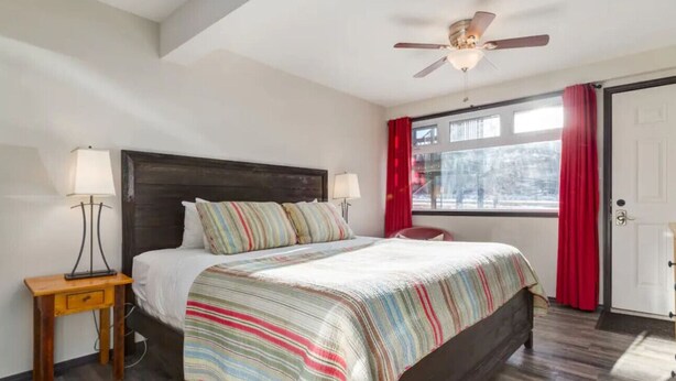 A bedroom with a bed, two bedside tables, a ceiling fan, a window with curtains, and a door.