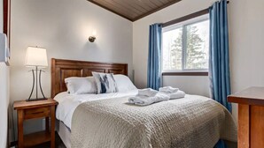 Ein ordentlich gemachteres Bett mit einer Steudecke, Kissen und gefalteten Handtüchern, einer Nachttischlampe und einem Fenster mit Vorhängen.