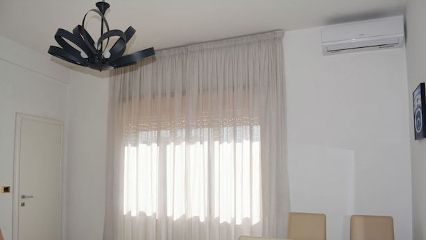 Een kamer met een plafondventilator, een witte airconditioner, een groot raam met dunne gordijnen, een beige bank en een klein wit tafeltje.