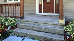 A wooden porch with a front door and windows, steps leading up to the entrance, and a small garden area with plants and a lantern.