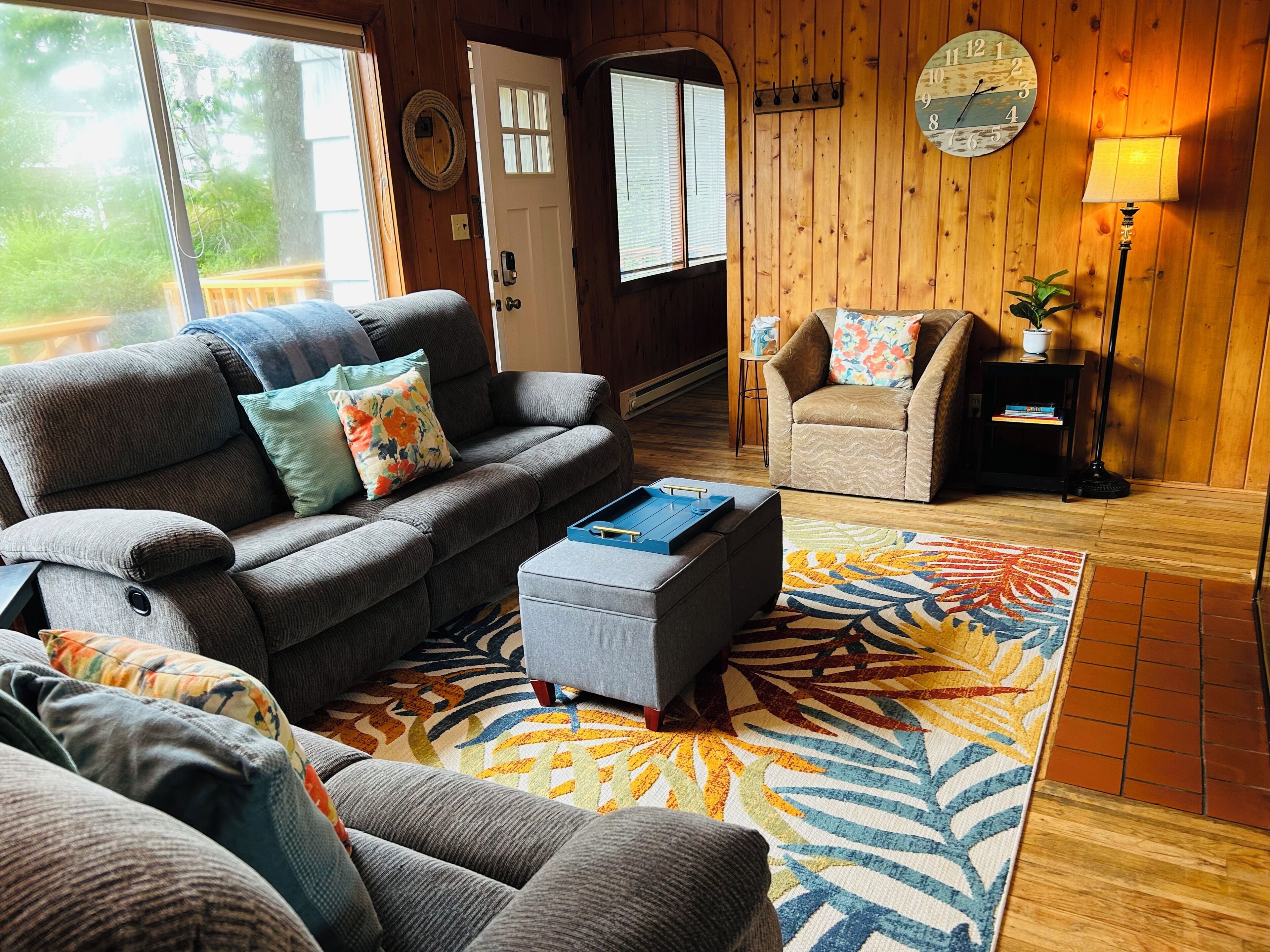 Ein gemütliches Wohnzimmer mit grauem Sofa, passendem Hocker, einem gemusterten Teppich, Holzboden, einer Uhr und einer Lampe.