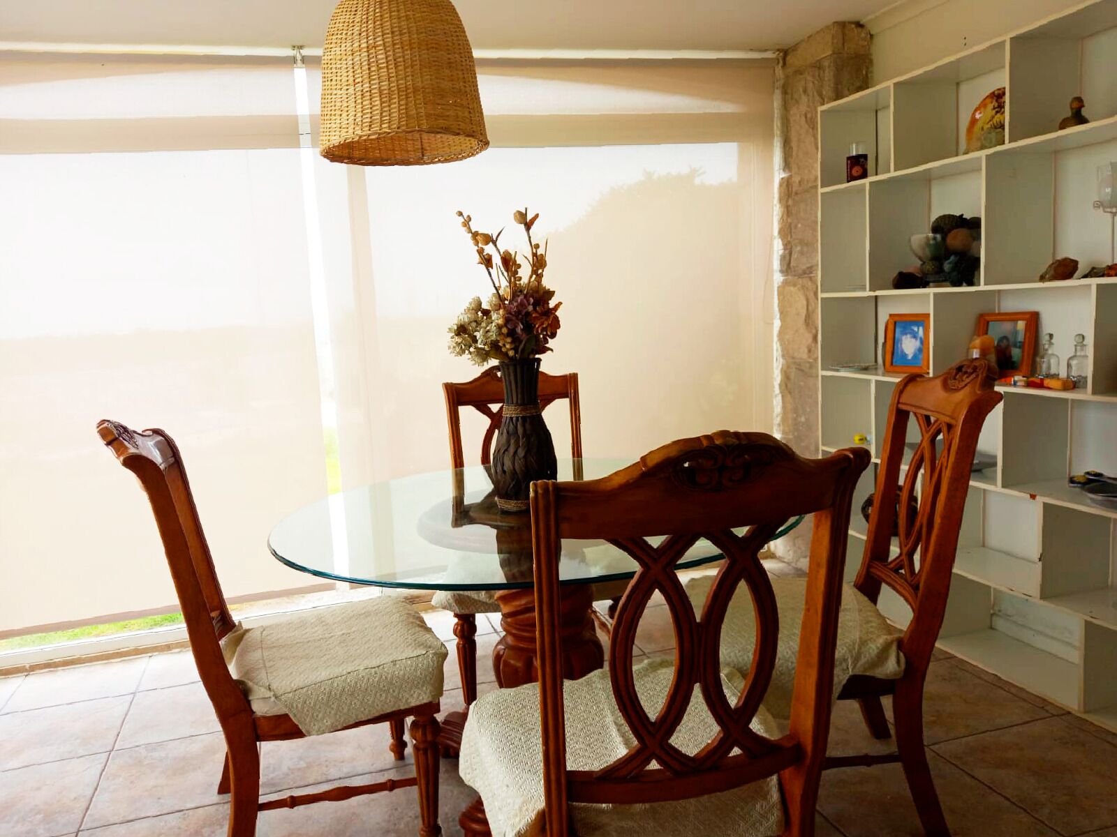 A dining area with a glass table, wooden chairs, a woven pendant light, and a vase with flowers.