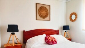 Un letto rifatto con lenzuola bianche e una testiera rossa, affiancato da due comodini con lampade, un quadro incorniciato sopra il letto e uno specchio rotondo sulla parete.