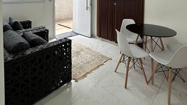A modern living room with a sofa, a dining table with chairs, a flat-screen TV mounted on a wooden panel, and a sliding glass door leading to an outdoor area. - CASA DA ALEGRIA (Praia Grande)