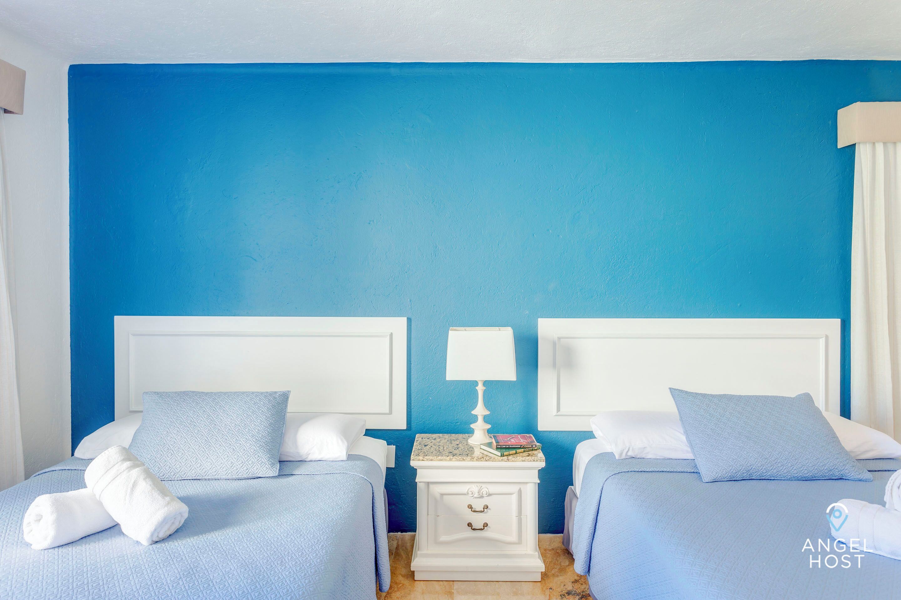 A hotel room with two beds, each with a blue bedspread and white pillows. There is a white nightstand with a lamp and books between the beds.