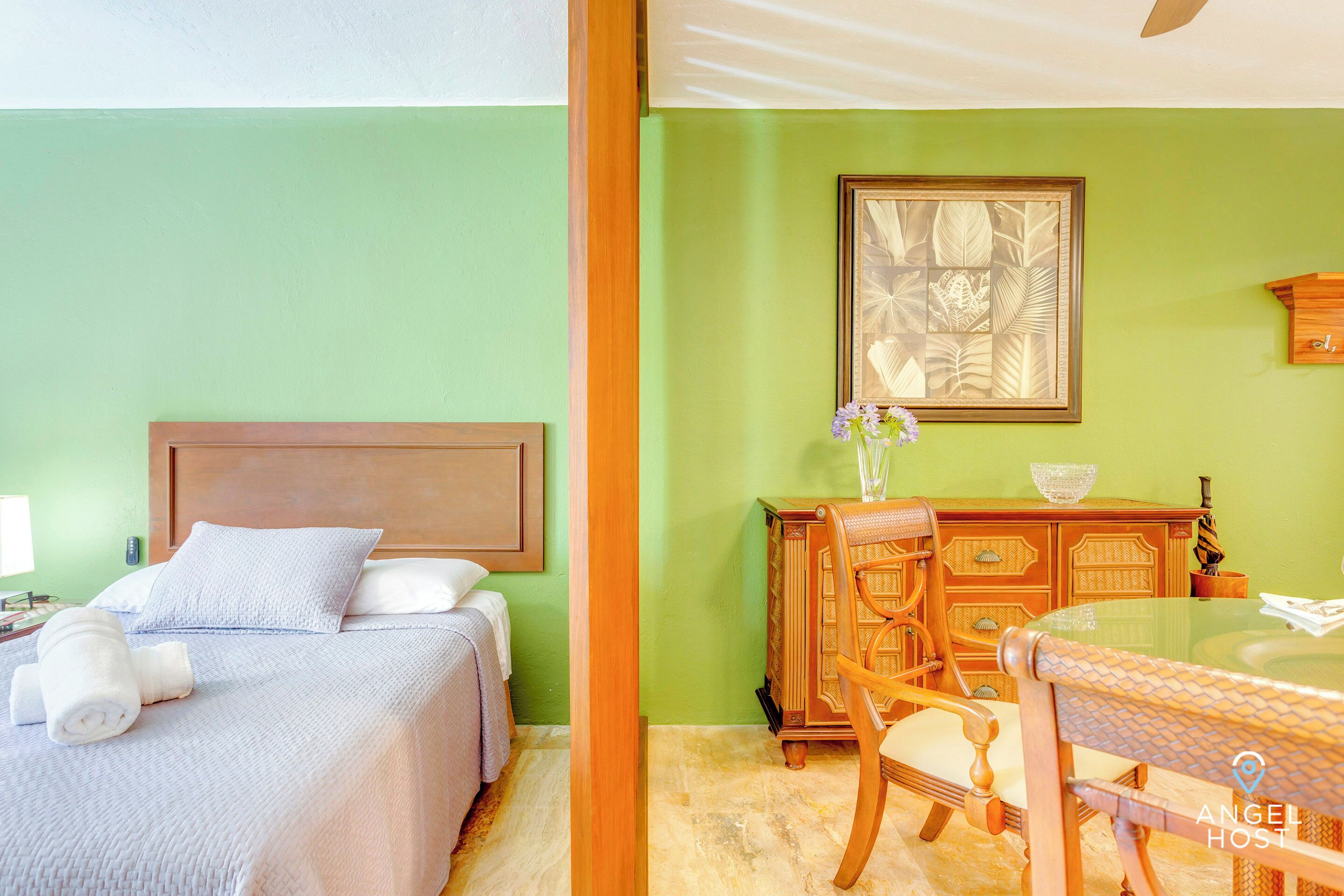 A hotel room with a bed, a wooden dresser, a dining table, and a framed artwork on the wall.