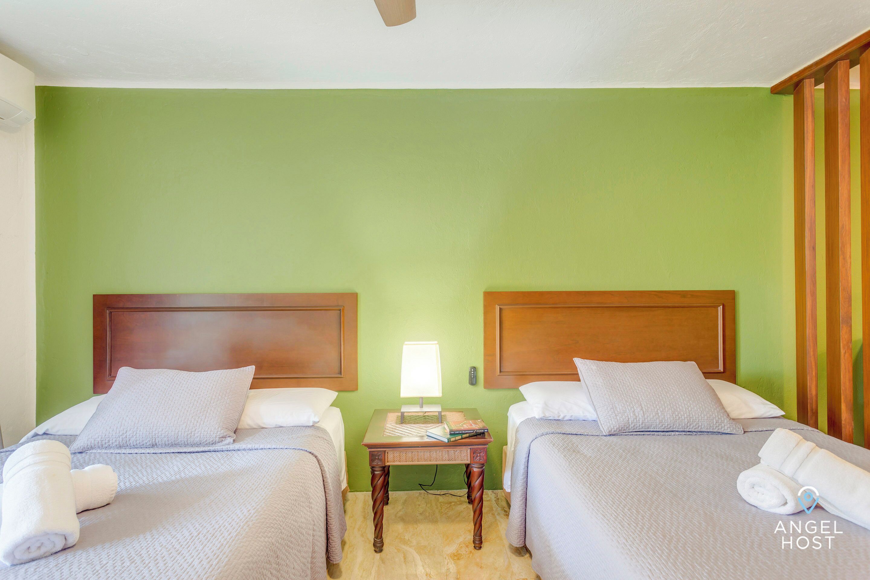 A hotel room with two beds, a nightstand with a lamp, and a green wall.