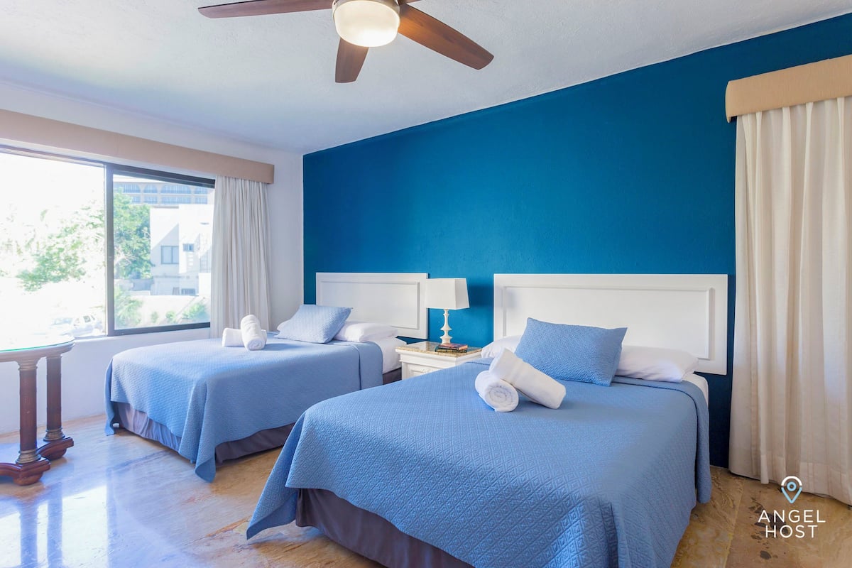 Une chambre d’hôtel avec deux lits, un ventilateur de plafond, une fenêtre avec des rideaux et une vue sur les bâtiments environnants.