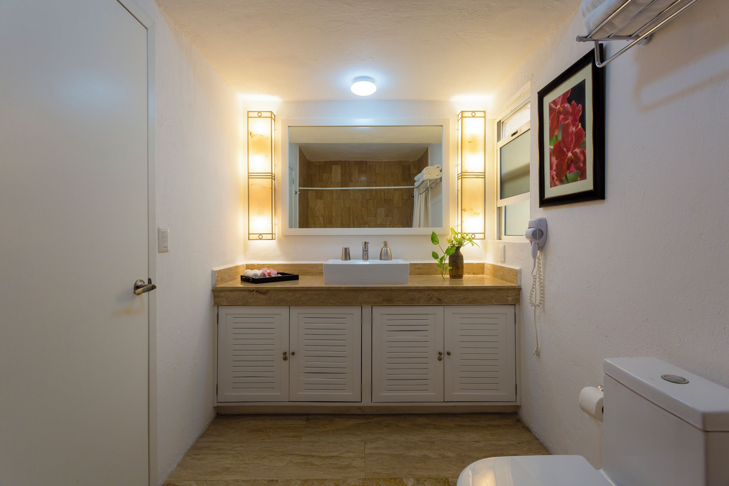 A bathroom with a wooden vanity, a white sink, a mirror with lights, a toilet, and a framed picture on the wall.