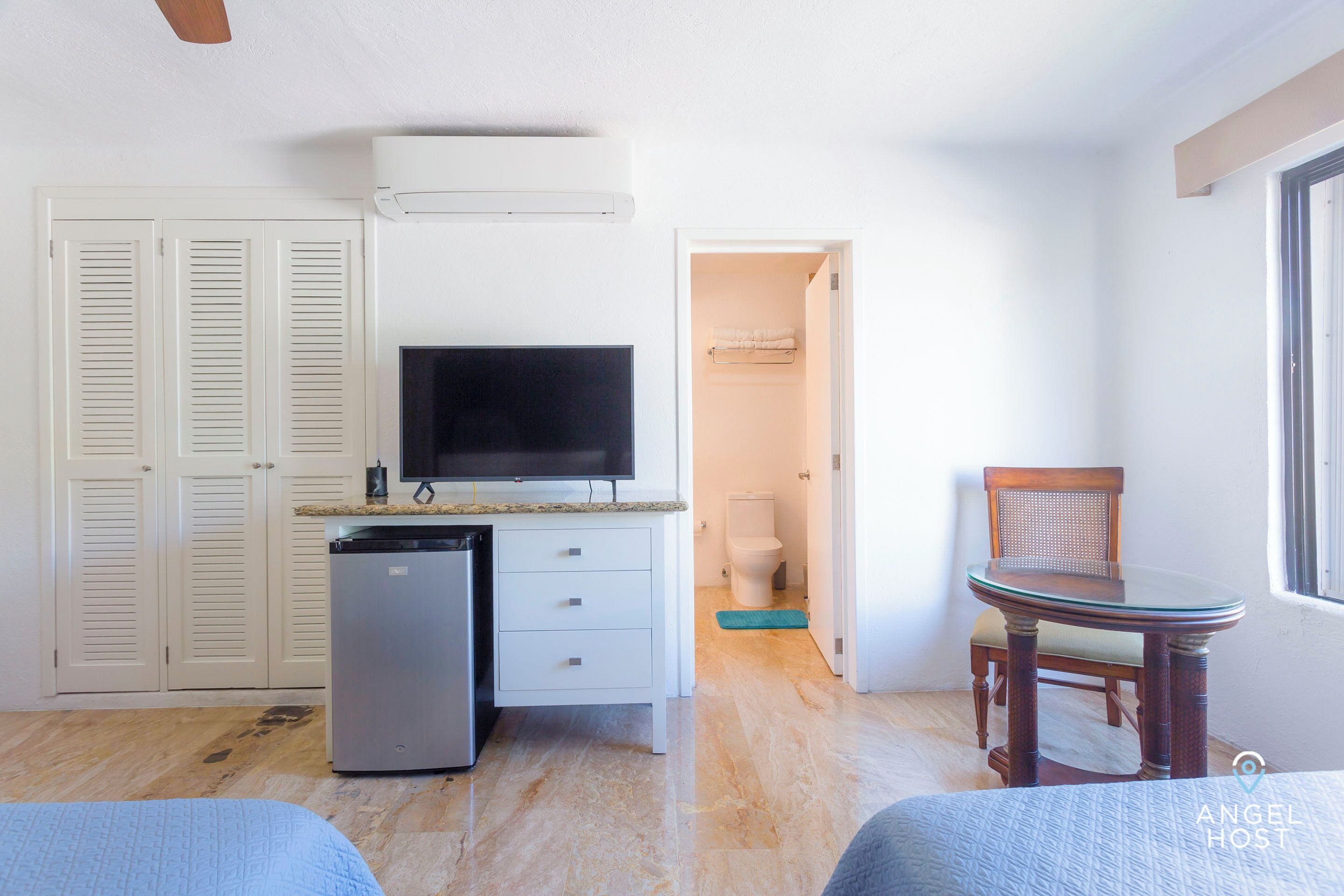 A compact room with a kitchenette, a flat-screen TV, a small refrigerator, a wooden chair, and a bed.