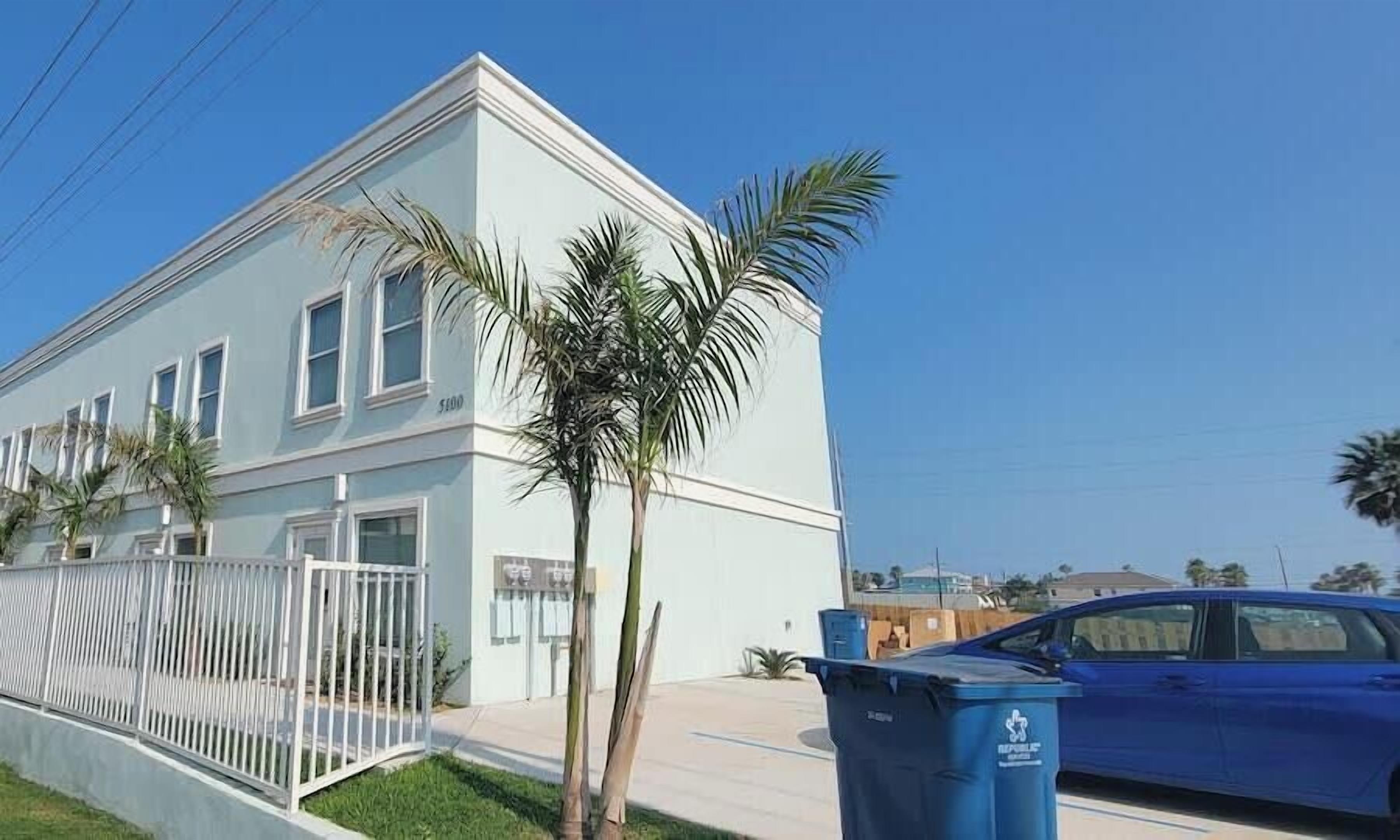 A light blue two-story building with a white fence, palm trees, and a parked blue car.