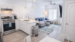 Cocina compacta con horno integrado, lavamanos y cafetera. En la sala de estar, hay un sofá gris con cojines azules, una mesa de comedor blanca y una alfombra pequeña.