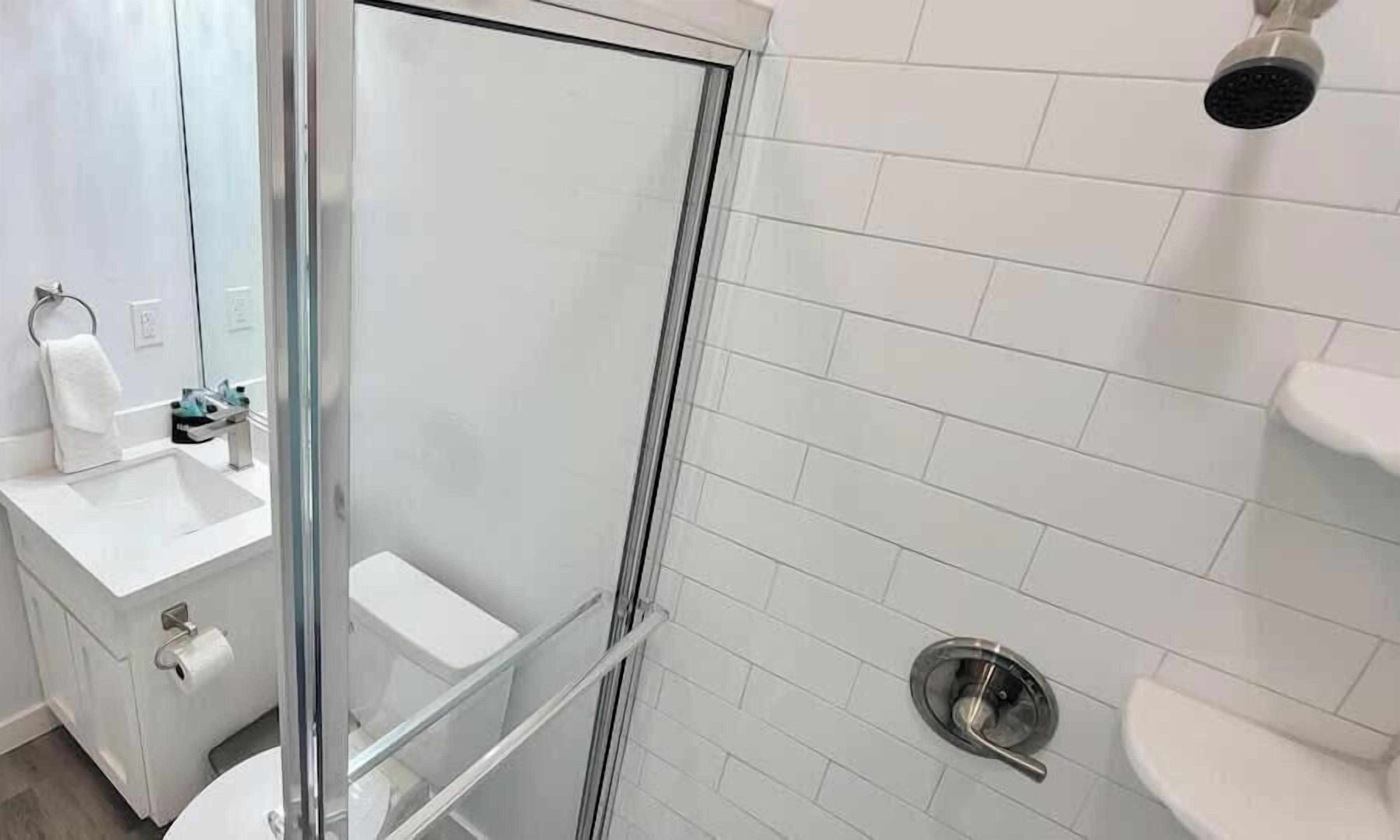 Un baño moderno con cabina de ducha de vidrio, azulejos blancos, un inodoro y un lavabo con artículos de aseo.