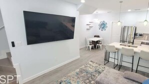 Un salon moderne doté d’un téléviseur à écran plat, d’un coin repas avec une table et des chaises, et d’une cuisine avec un bar.