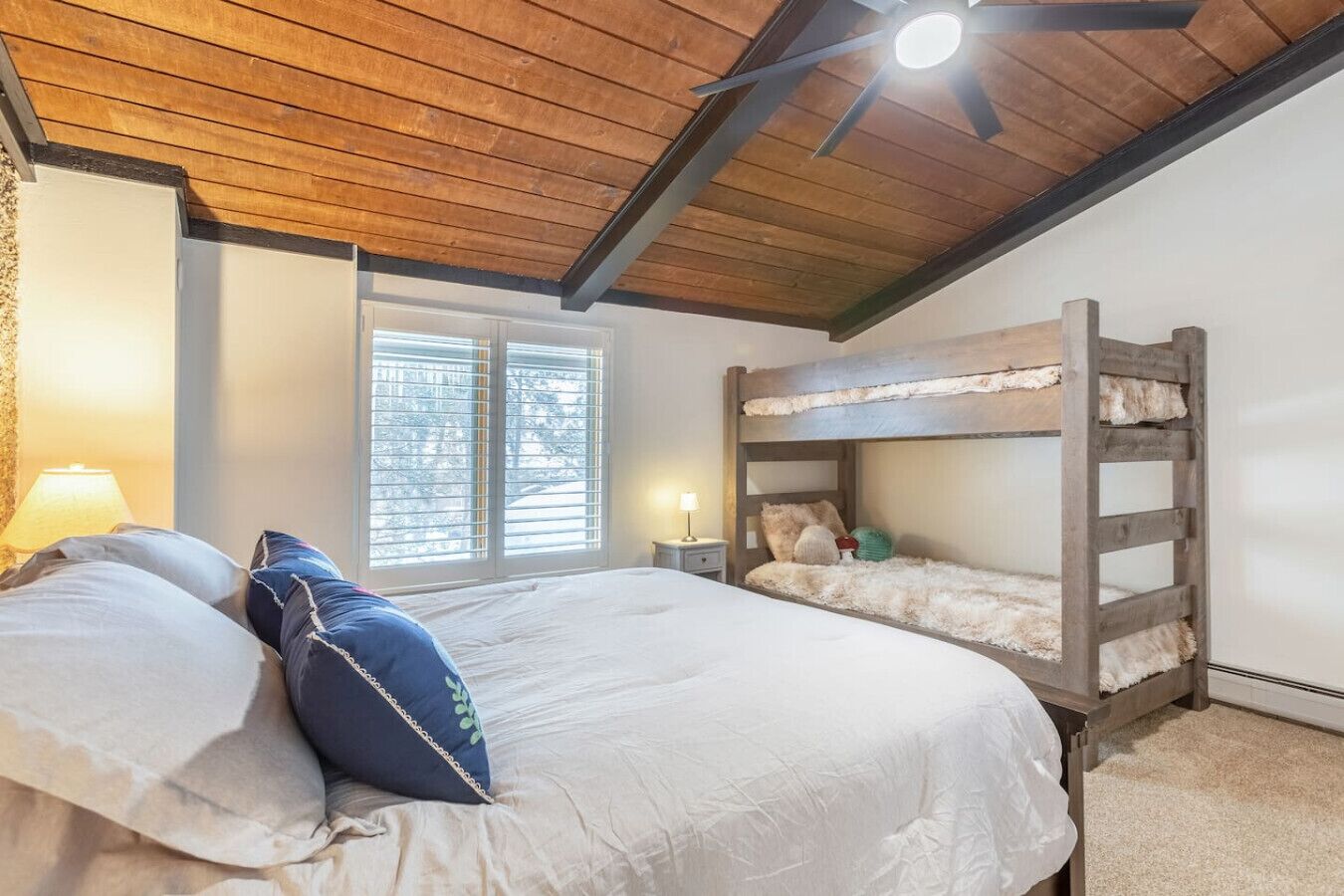 A bedroom with a bunk bed, a large bed with pillows, a ceiling fan, and a window with blinds.
