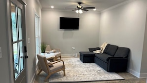 A living room with a ceiling fan, a flat-screen TV mounted on the wall, a black sectional sofa, a beige armchair, and a patterned rug. - The Gray Cottage on Brownlee! (Calhoun)